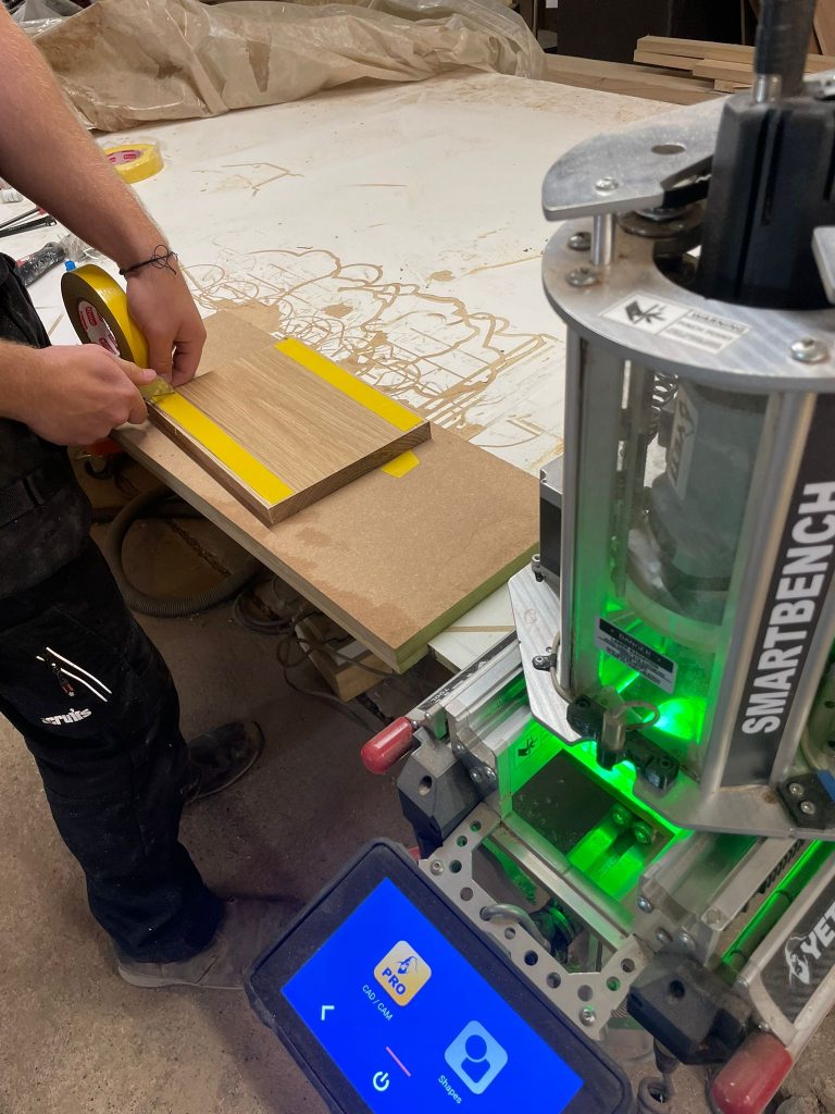 A CNC operative securing a solid oak board onto the router bed with double-sided tape before precision engraving — preparation for CNC machining in Cambridgeshire.