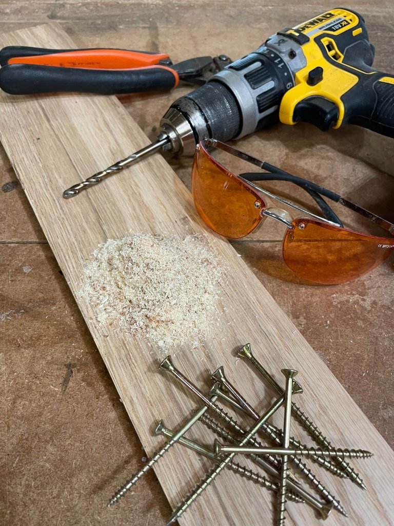 DeWalt drill, safety glasses and screws arranged on a wooden board with sawdust — workshop tools used by KMP Carpentry & Joinery in Cambridgeshire.