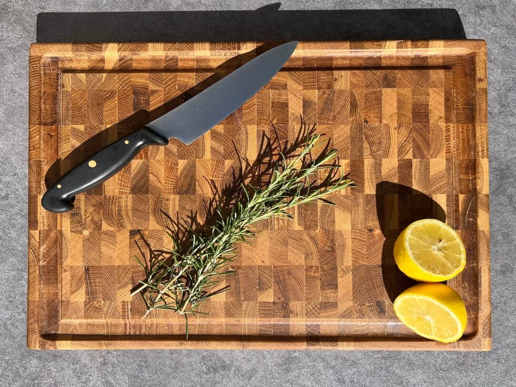 Overhead view of a personalised end grain oak chopping board with juice groove, kitchen knife, rosemary sprig and sliced lemon.