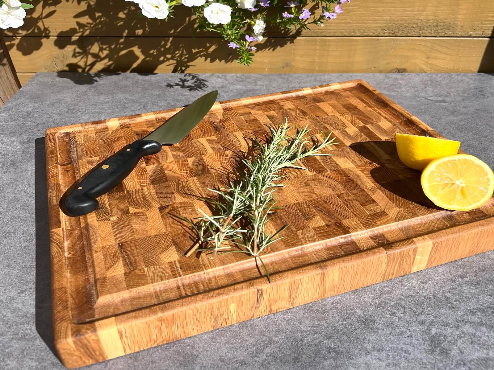 Angled view of a personalised oak end grain chopping board with knife, rosemary and lemon outdoors.