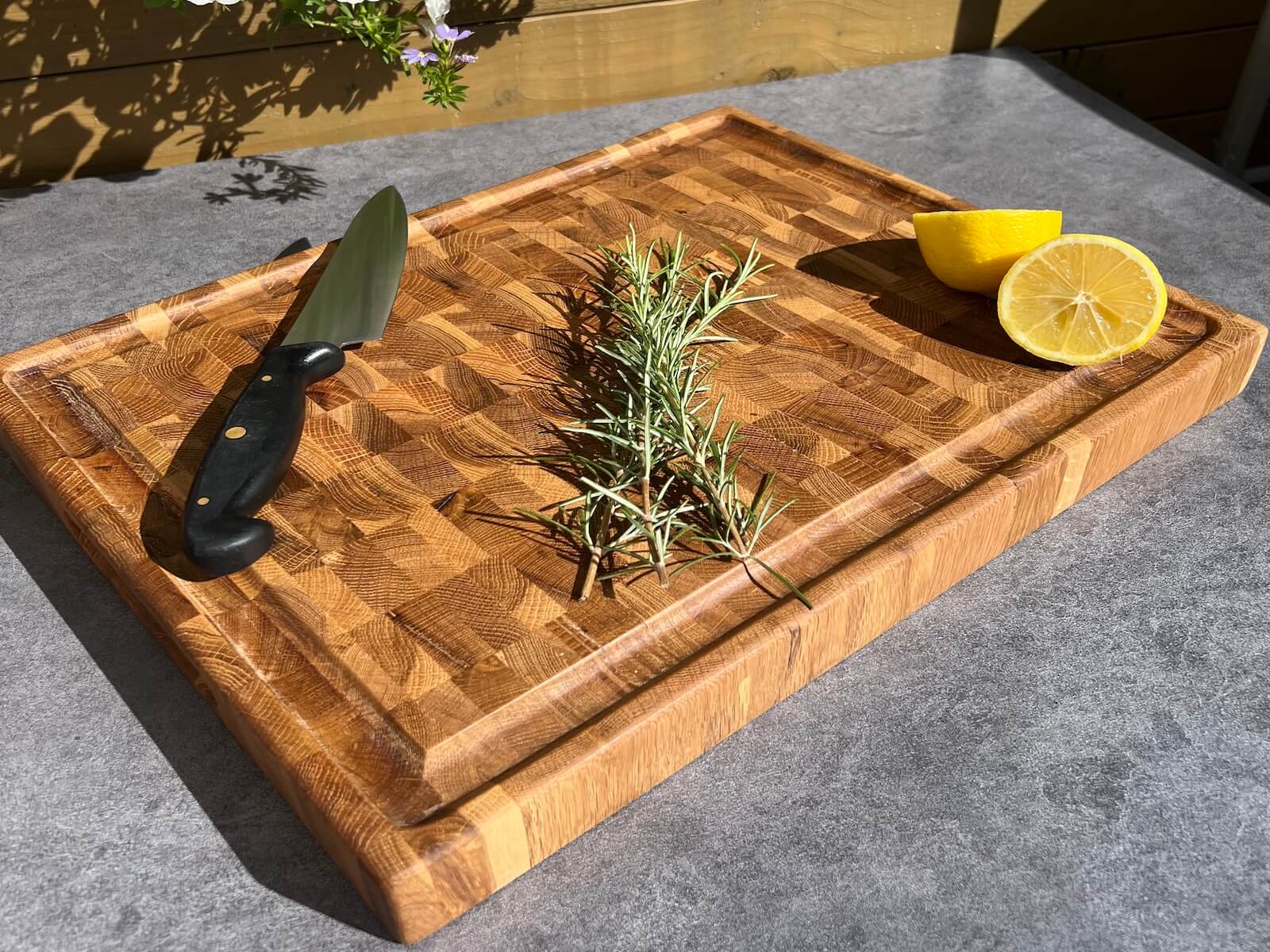 Handmade oak end grain chopping board with engraved detailing, knife, herbs and lemon displayed outdoors.