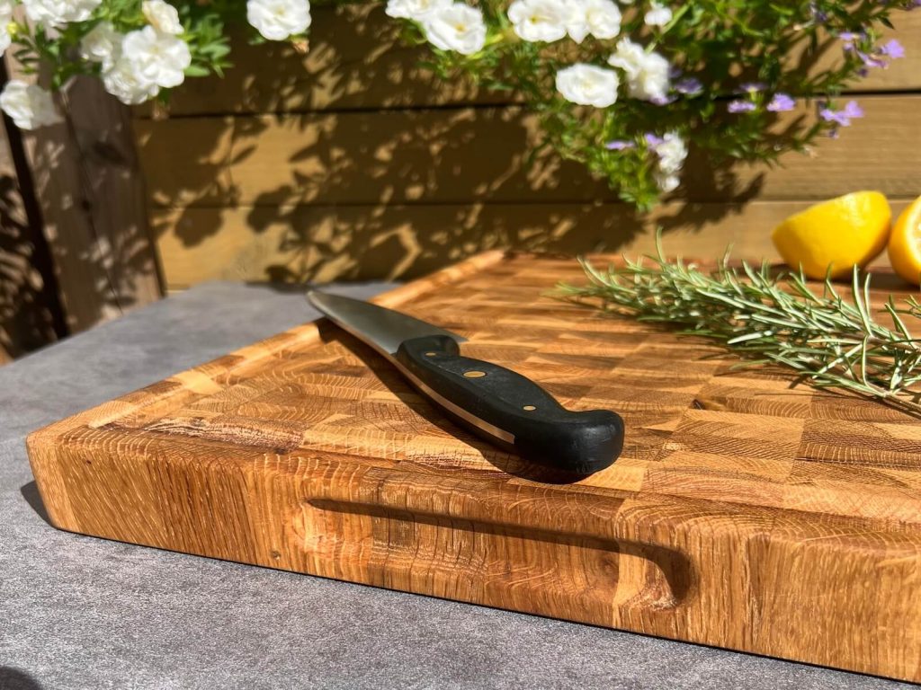 Close-up of a handmade oak end grain chopping board with side handles, knife, rosemary and lemon.
