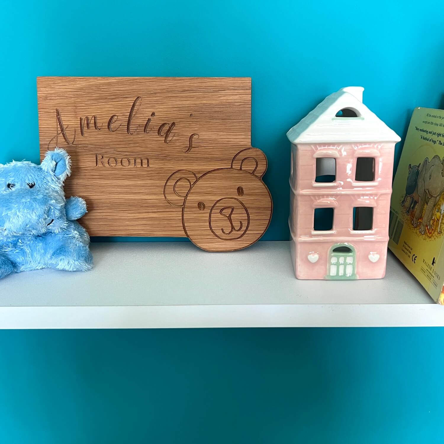 Personalised oak children’s bedroom sign engraved with teddy bear design, styled on a shelf with toys.