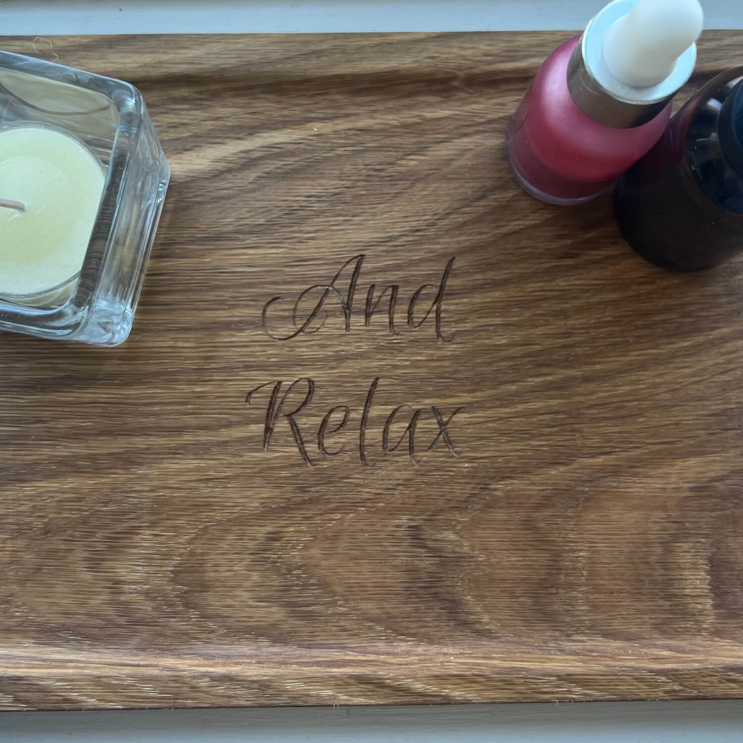 Close-up of oak display tray engraved with “And Relax,” styled with candle and skincare products.
