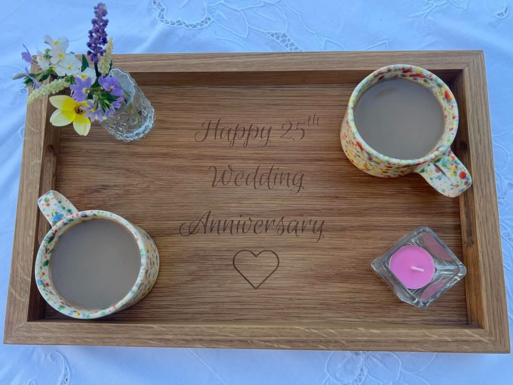 Handmade oak serving tray engraved with “Happy 25th Wedding Anniversary” surrounded by mugs of tea, flowers and a candle.