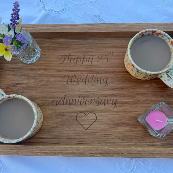 Handmade oak serving tray engraved with “Happy 25th Wedding Anniversary” surrounded by mugs of tea, flowers and a candle.