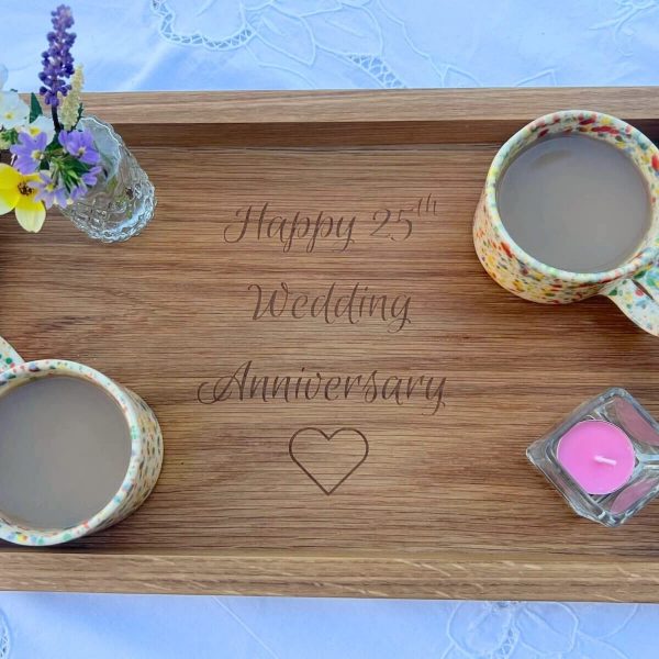 Handmade oak serving tray engraved with “Happy 25th Wedding Anniversary” surrounded by mugs of tea, flowers and a candle.