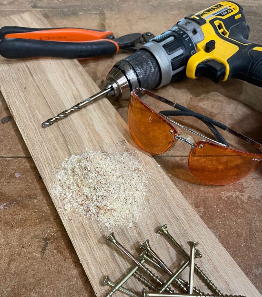 DeWalt drill, safety glasses and screws arranged on a wooden board with sawdust — workshop tools used by KMP Carpentry & Joinery in Cambridgeshire.