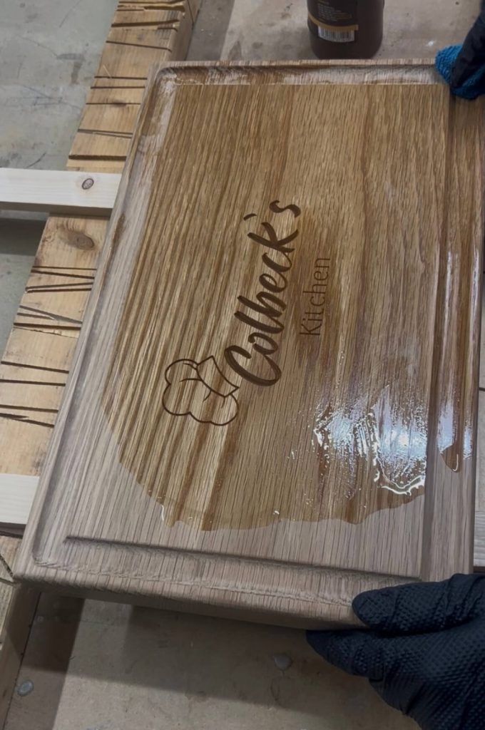 A pair of gloved hands applying food-safe oil to a completed personalised oak chopping board with engraved text.