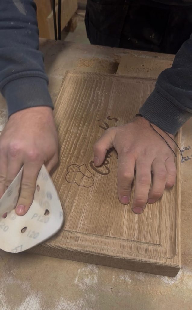 Hand sanding a personalised solid oak chopping board at KMP Joinery to smooth the engraved design and prepare it for finishing.