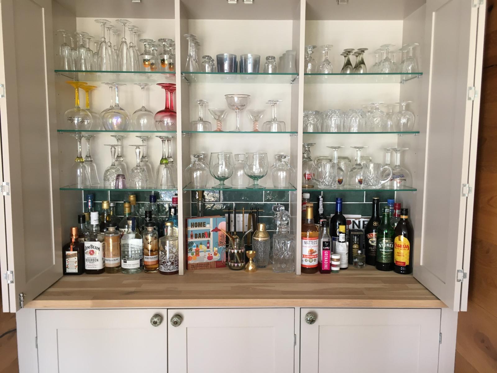 Bespoke fitted home bar with painted cupboards, glass display shelves and timber worktop in Cambridgeshire.